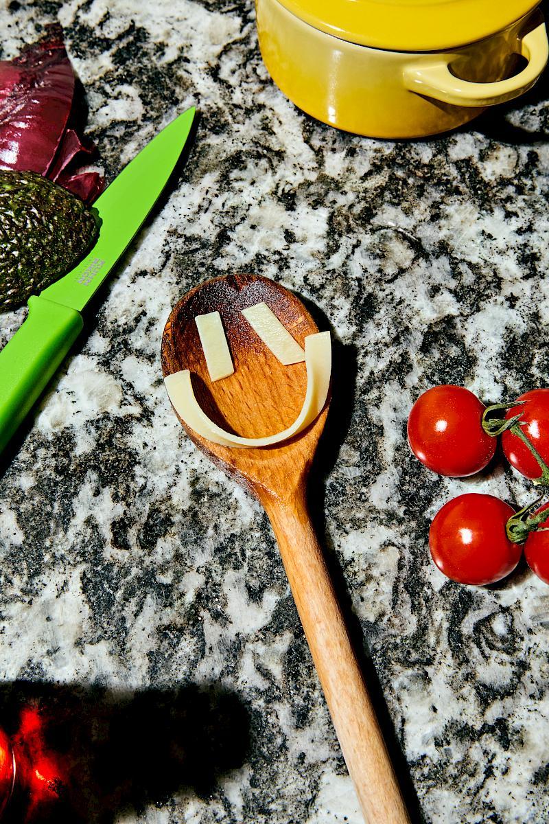 Smiling wooden spoon surrounded by cherry tomatoes, avocado, onion skin and a yellow Le Creuset mini-Cocotte