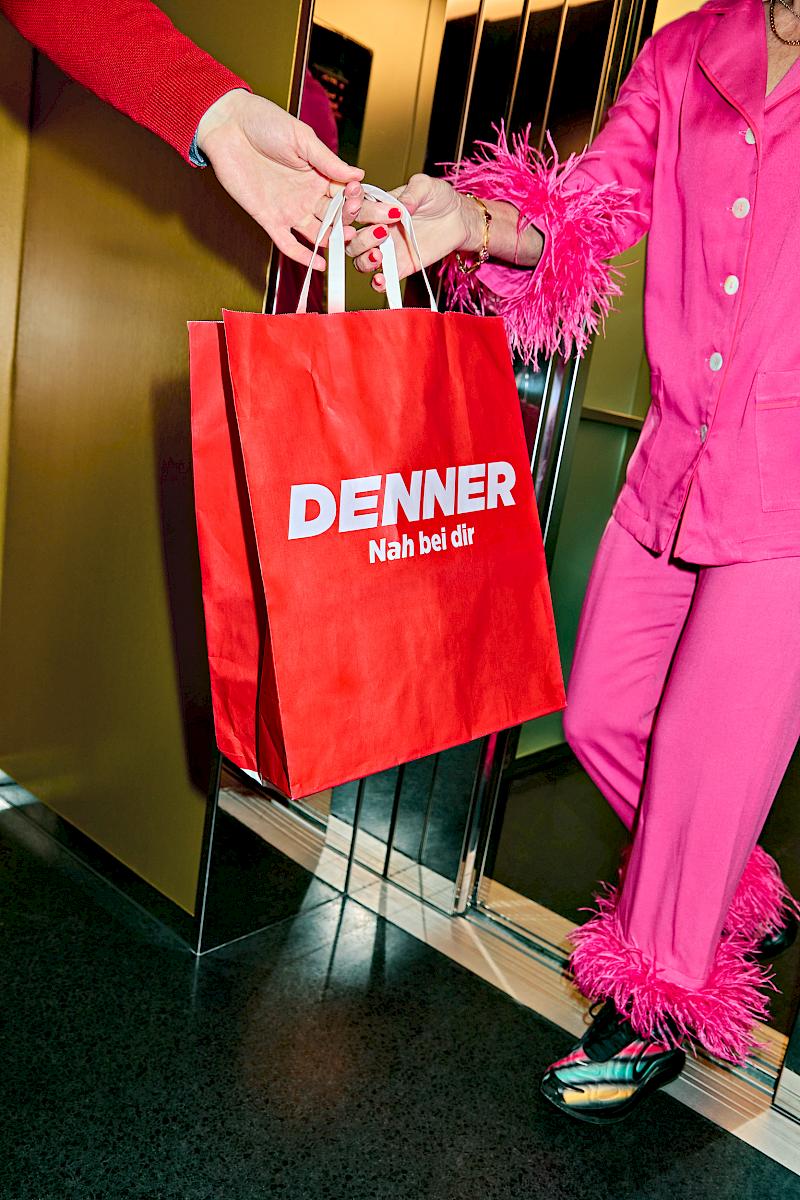 A man hands over a Denner groceries bag to a lady in a hot pink party pyjama with detachable feathers