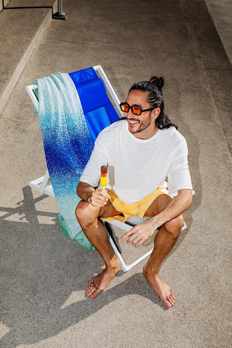 A man sitting on a SULA x Micasa deck chair outdoors, holding a colourful popsicle. He is wearing sunglasses and a white t-shirt, with yellow shorts
